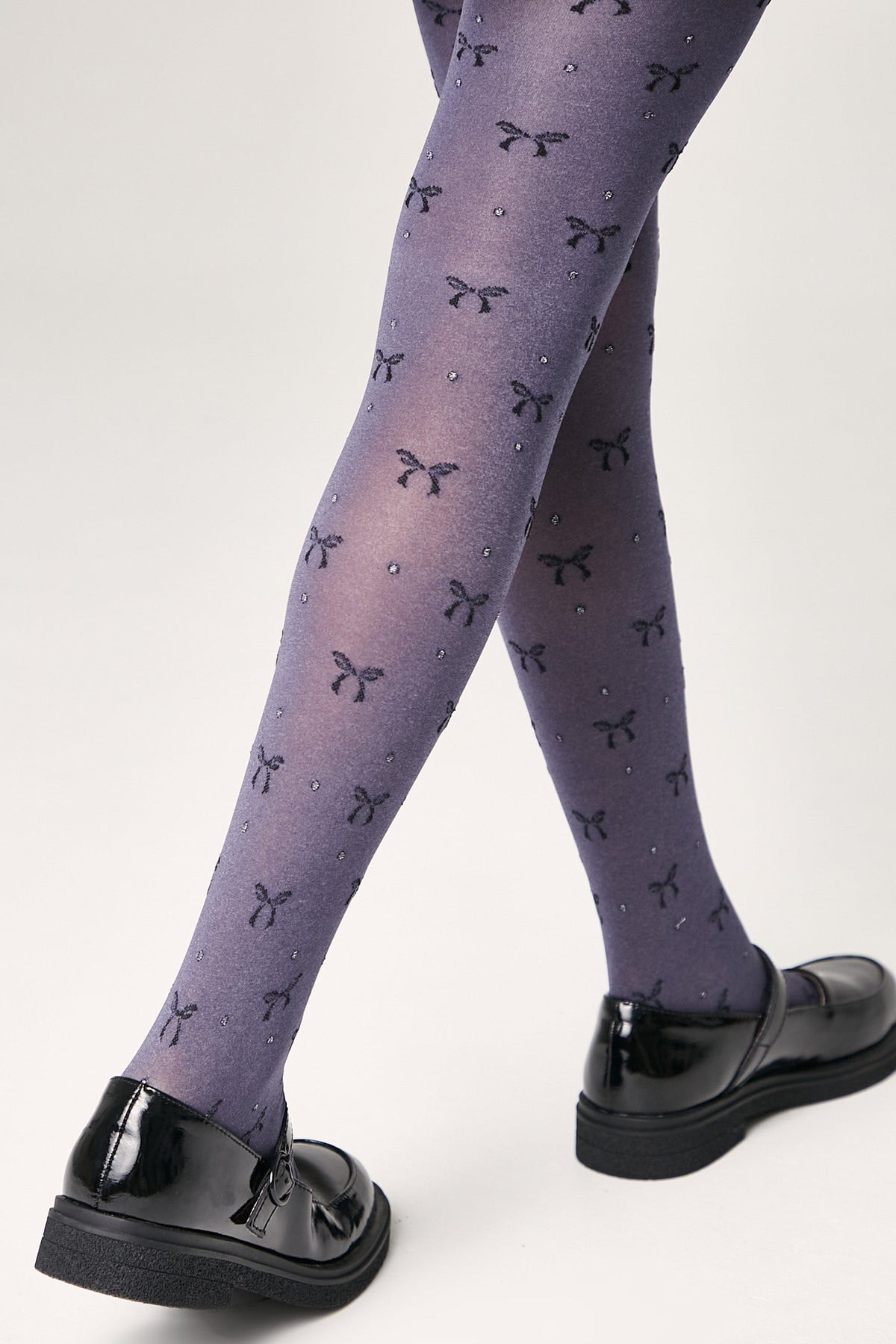 A pair of purple semi-sheer tights featuring a repetitive pattern of small dark bows and tiny sparkling accents, worn with black patent leather mary jane shoes.