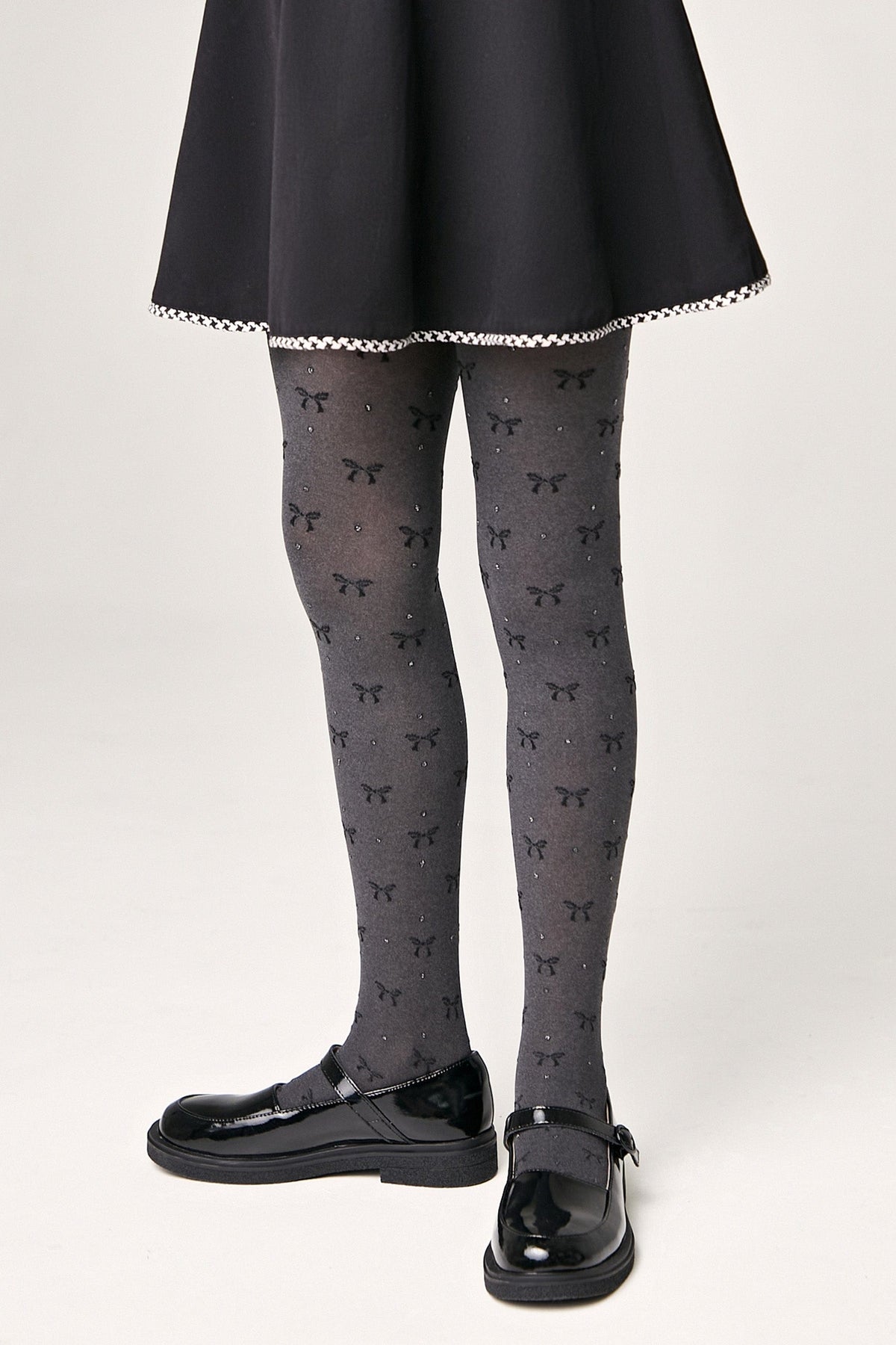 A person wears grey patterned tights featuring small black bows and subtle sparkles, paired with a black skirt and shiny black buckled Mary Jane shoes.