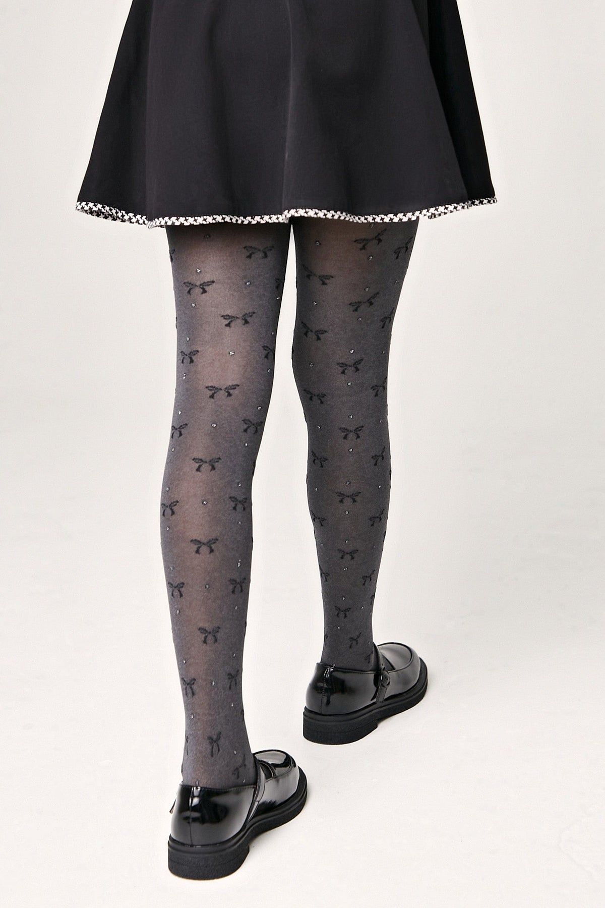 Gray sheer tights decorated with a repeating pattern of small black bows and subtle sparkling rhinestones worn with a black skirt and black loafer-style shoes.