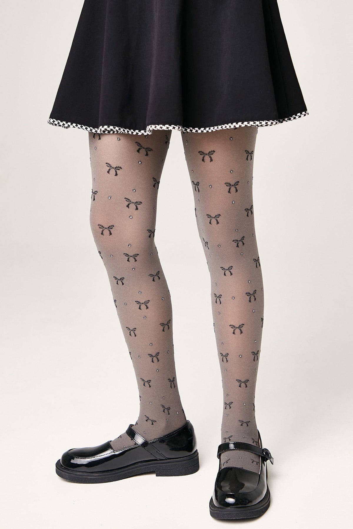 Sheer grey tights patterned with small black bows and sparkling rhinestones are worn by a person dressed in a black skirt and black mary jane shoes.