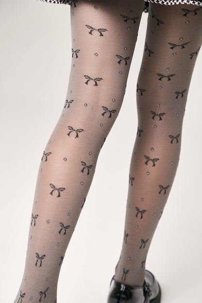 Sheer grey tights patterned with a repeating print of small black bows and tiny sparkling dots, worn with black shoes.