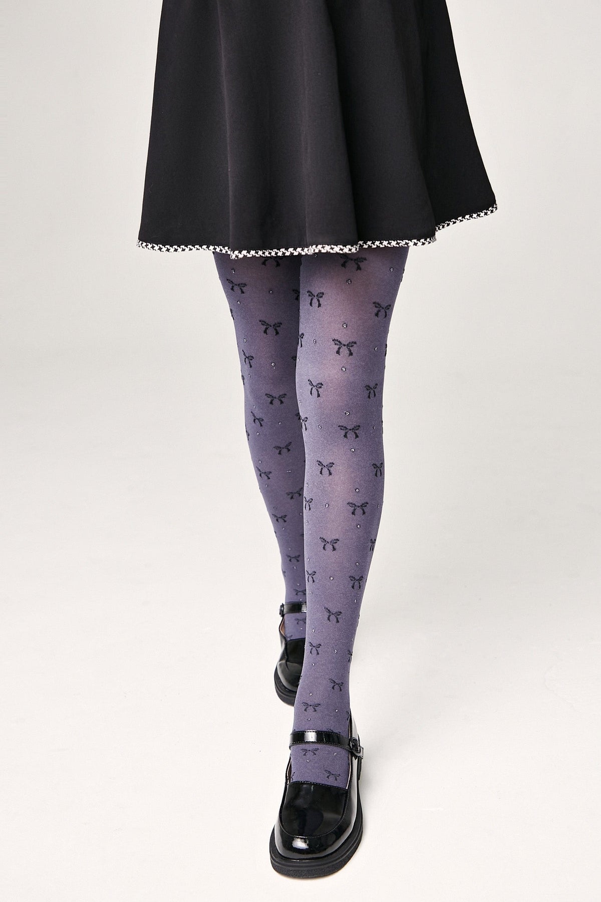 A person wears grey tights patterned with black bows and small sparkles, paired with a short black skirt and black mary jane shoes.