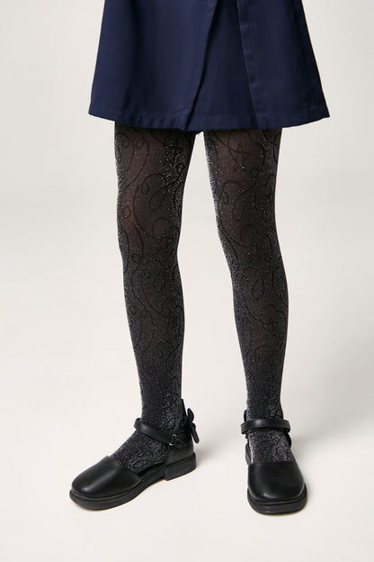 A pair of black glittery tights featuring a subtle swirling pattern worn under a navy blue skirt with black buckled flat shoes.