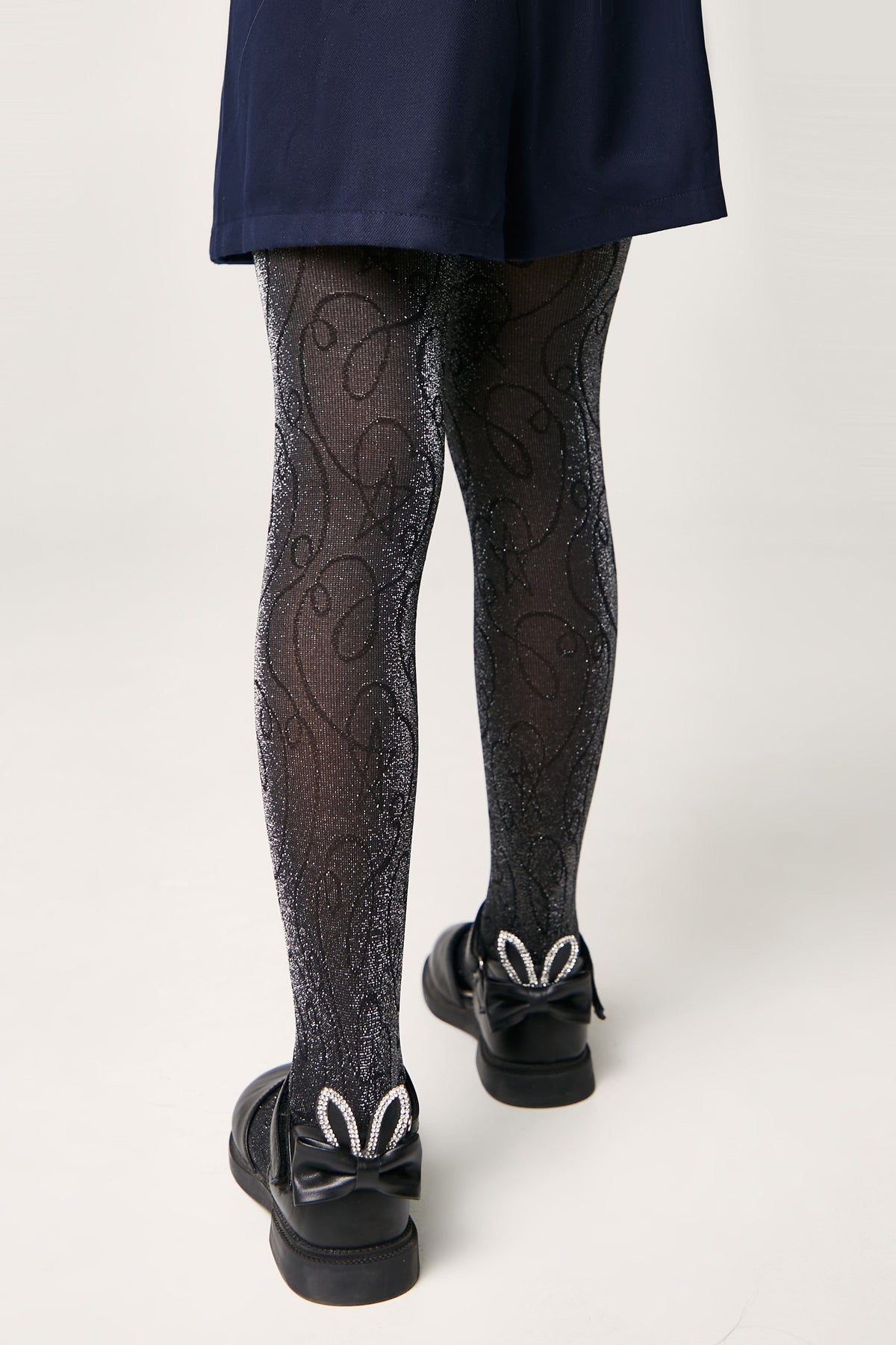 Black tights with a glittery swirling pattern worn with black shoes featuring a bow and decorative rabbit ears on the heel.