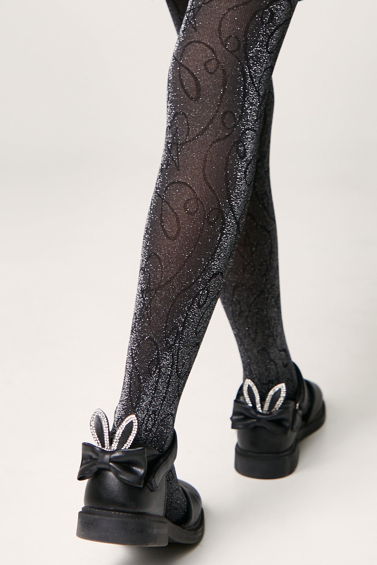 Black glittery tights with a swirling line pattern worn with black shoes featuring a bow and crystal-embellished rabbit ears on the heel.