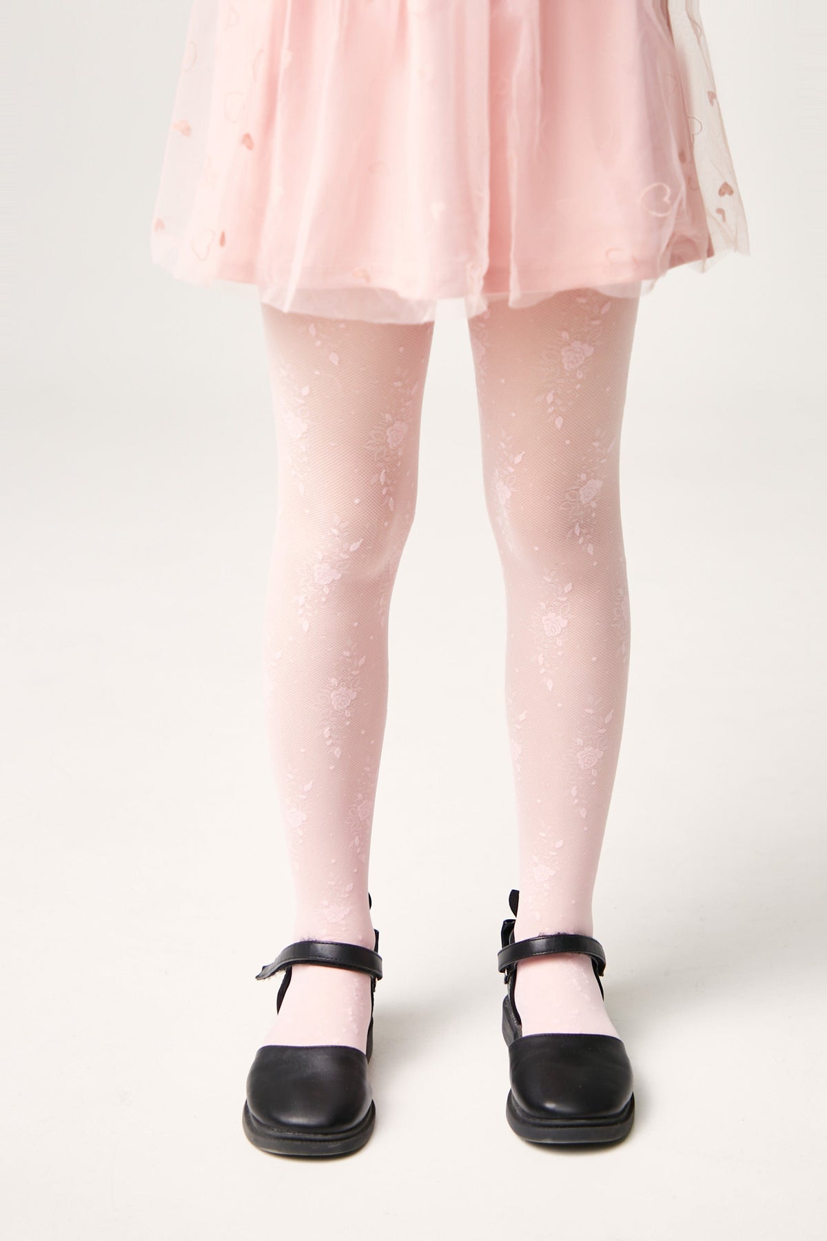 A pair of pale pink tights with a delicate floral pattern worn with a light pink skirt and black mary jane shoes against a plain white background.