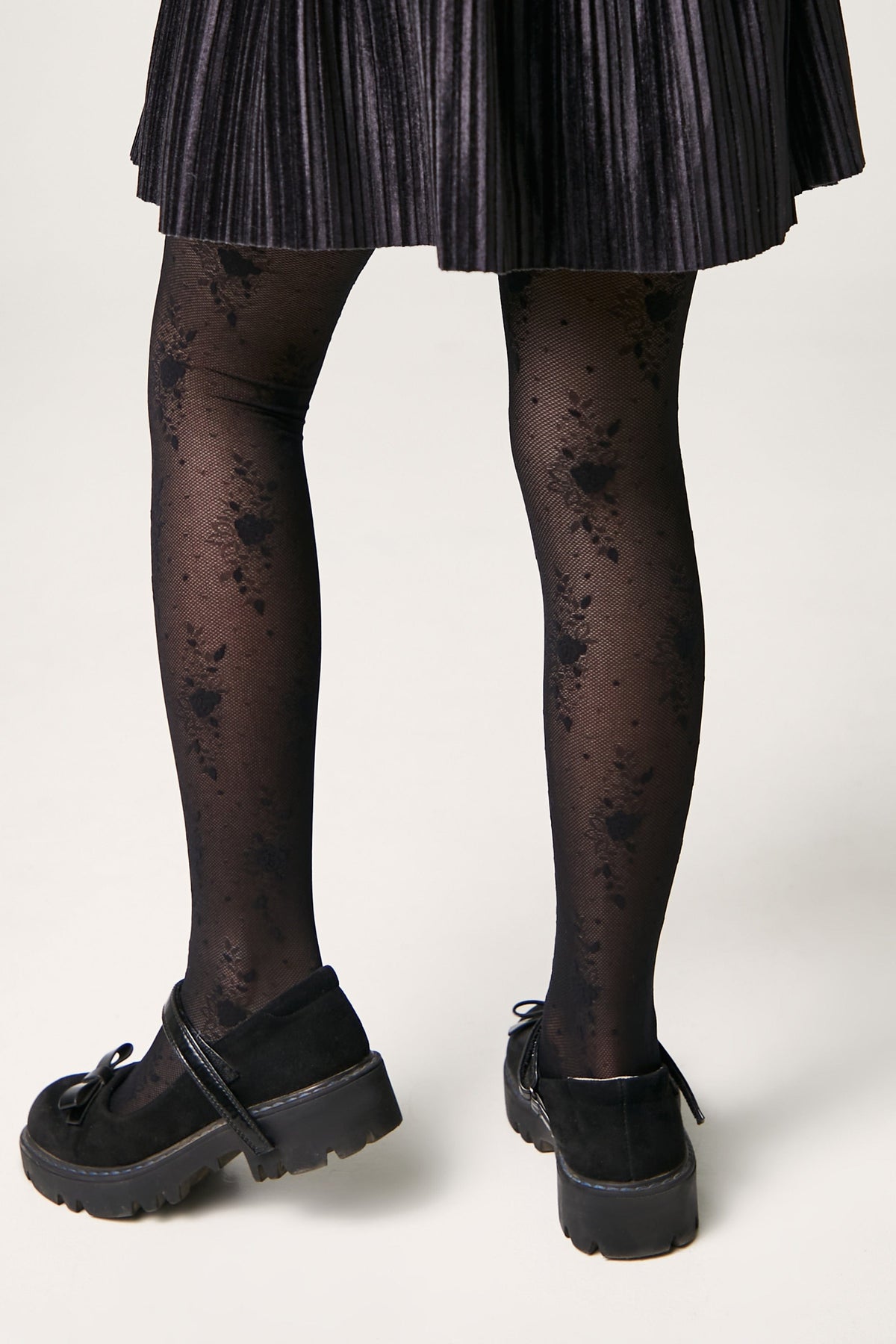 Black tights with a delicate floral lace pattern worn with black velvet Mary Jane shoes and a dark pleated skirt.