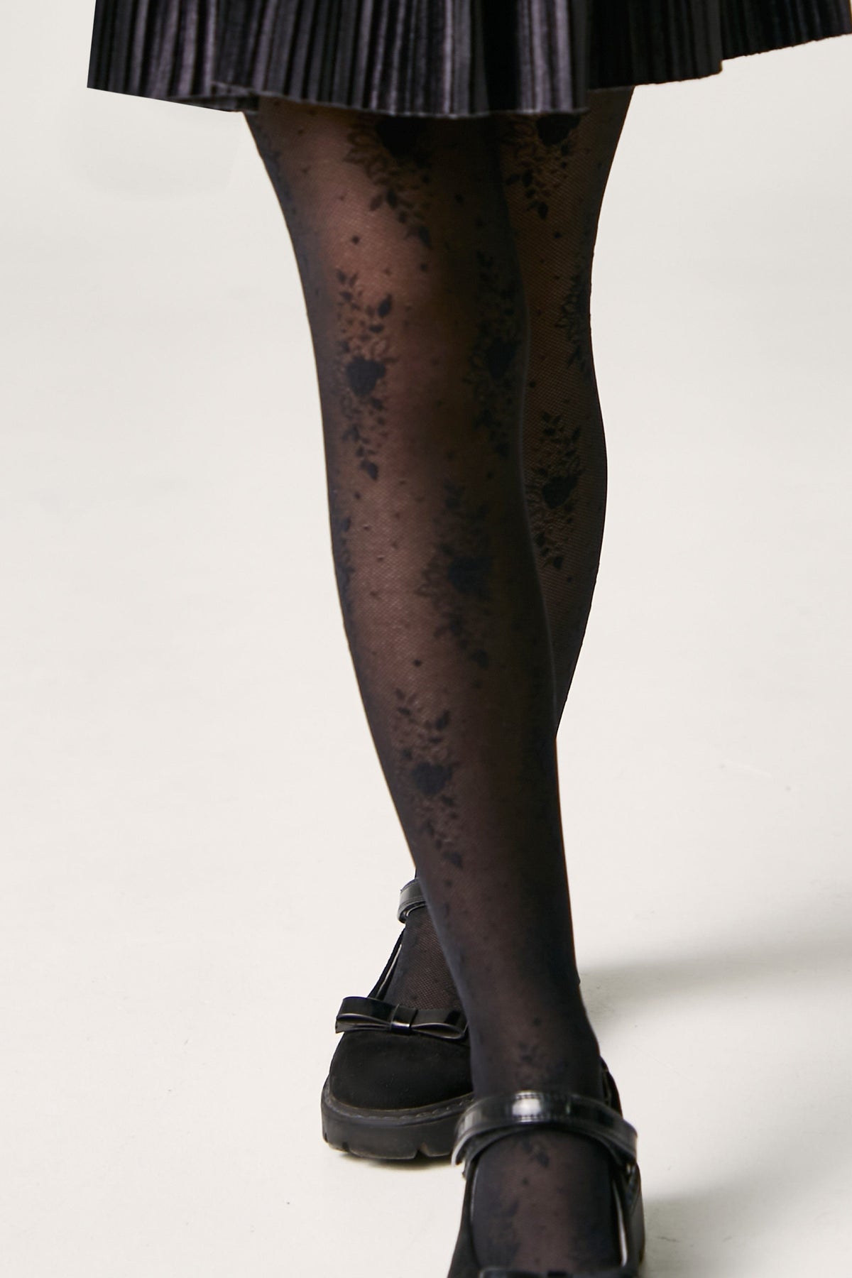 Sheer black patterned tights worn with black Mary Jane shoes and a black pleated skirt.