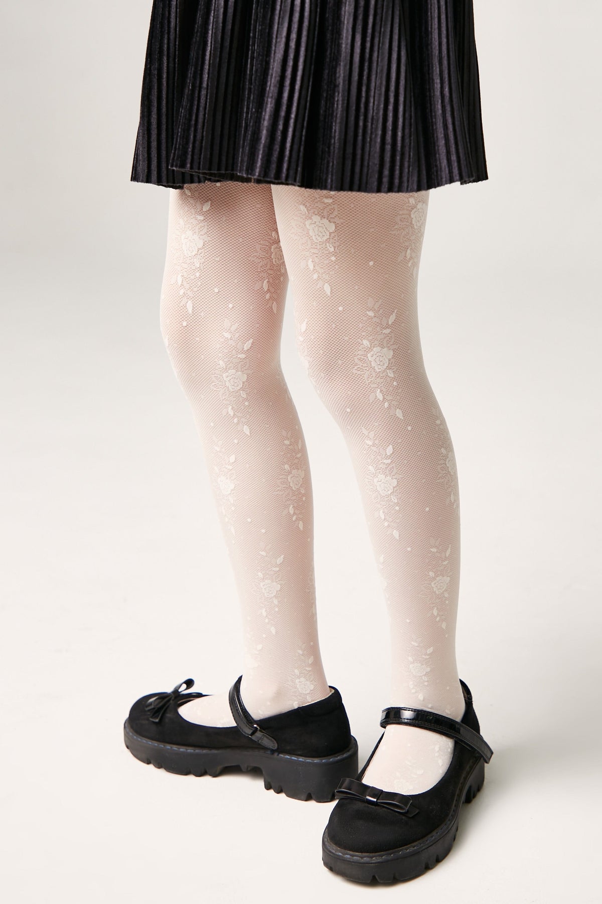 White tights featuring a delicate floral lace pattern worn with a black pleated skirt and black platform Mary Jane shoes.
