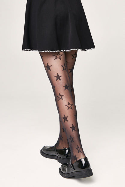 Sheer black tights patterned with various sized black stars are worn by a person dressed in a black skirt and glossy black flat shoes against a plain off-white background.