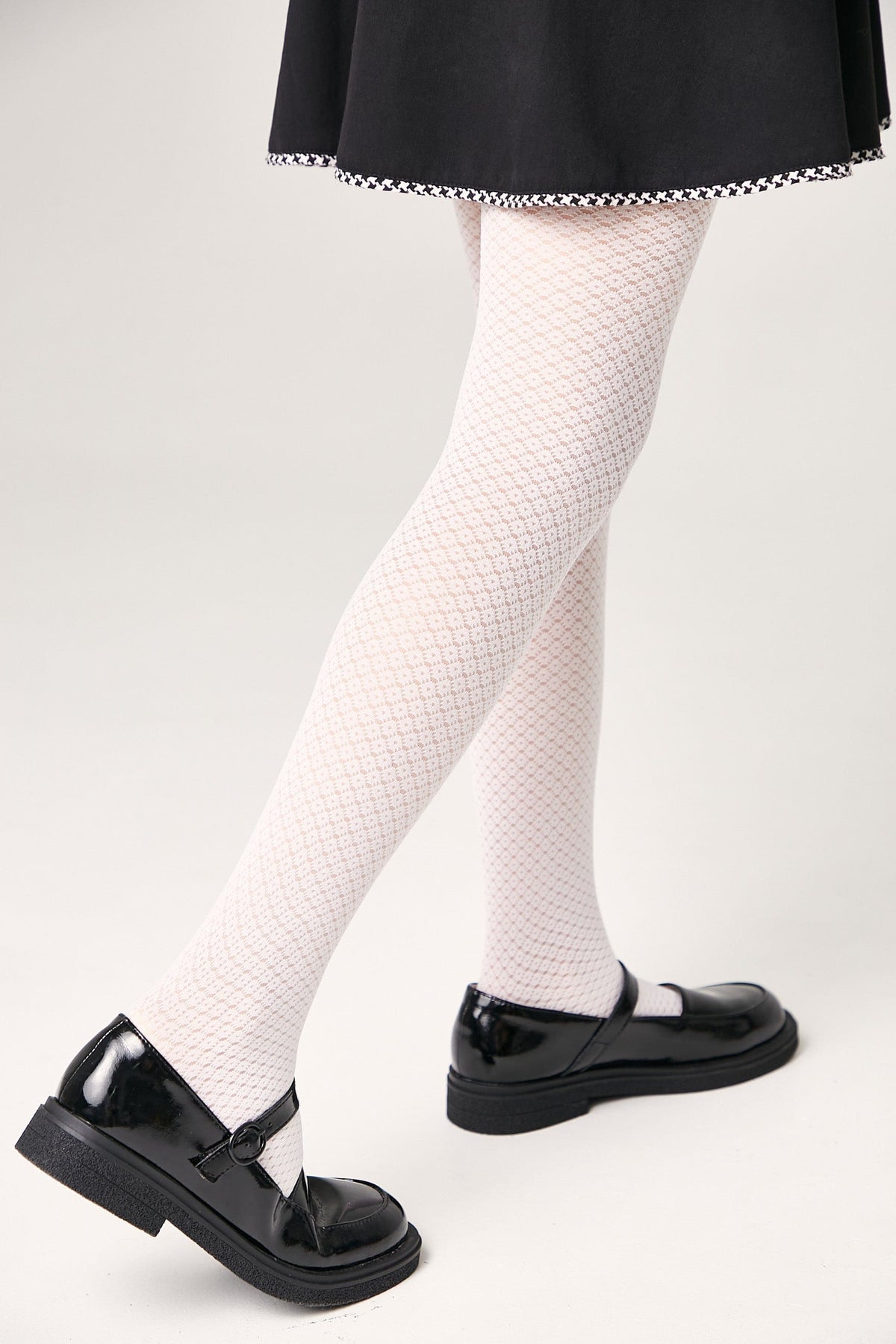 White textured tights are worn with a black skirt and glossy black Mary Jane shoes against a light gray background.