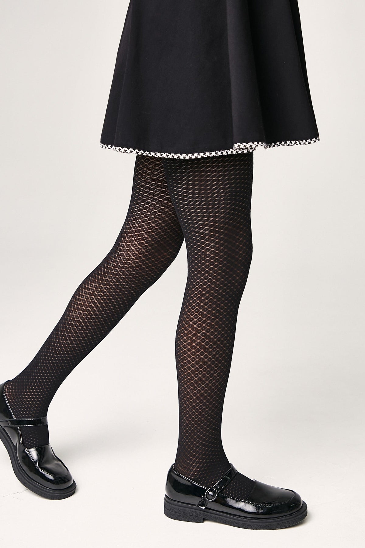 Black patterned tights worn with a black flared skirt featuring a houndstooth hem and black patent leather Mary Jane shoes against a white background.