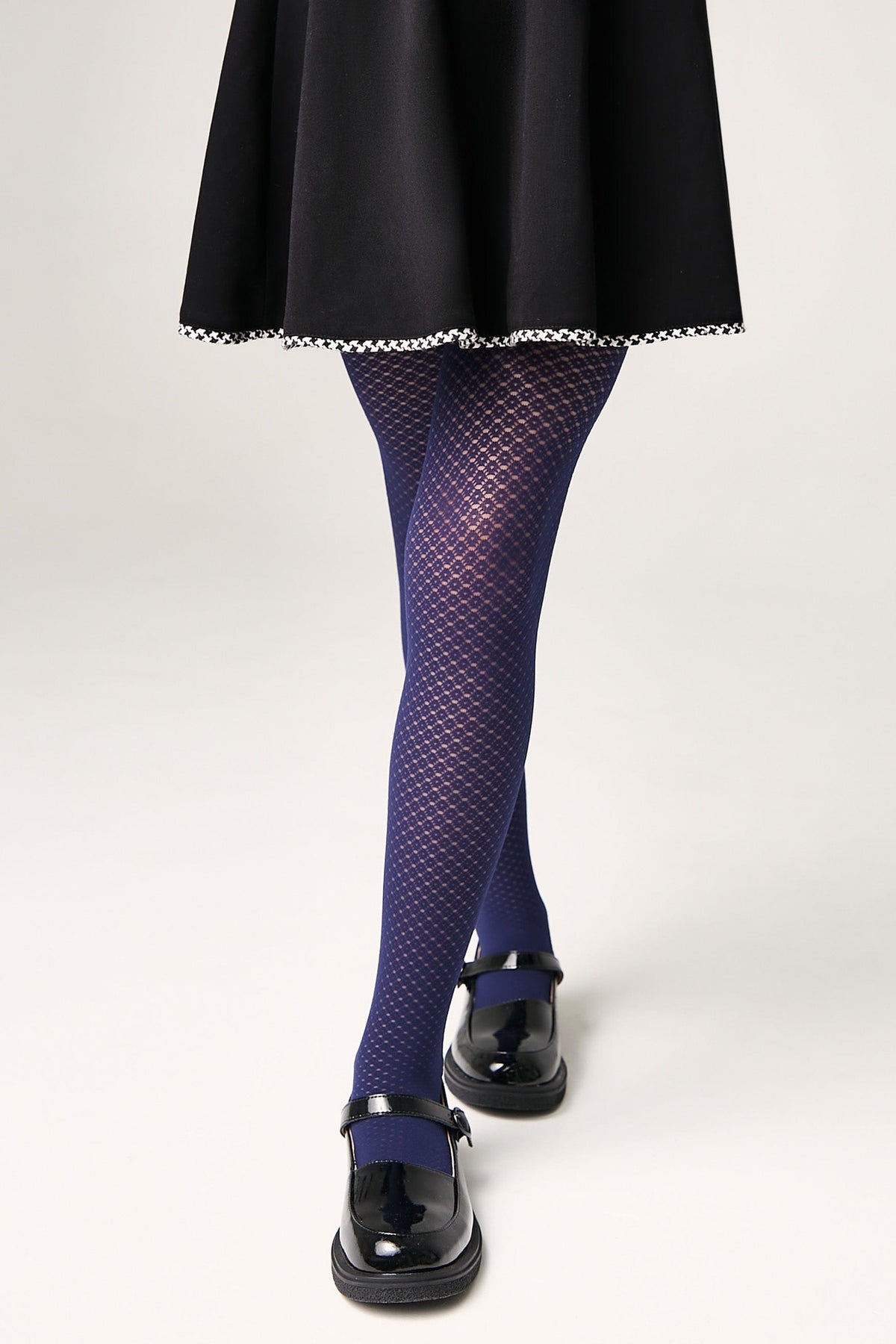 A person wears navy blue textured tights with a subtle diamond pattern, paired with a black skirt with white trim and black patent leather Mary Jane shoes against a plain white background.