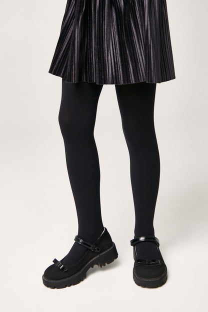 Black Mary Jane style shoes featuring a bow detail on the toe, chunky lug soles, and adjustable ankle straps worn with a pleated black skirt and opaque tights.