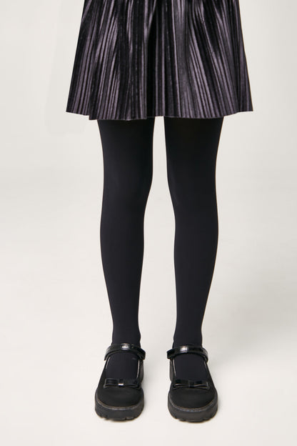 A person wears a dark grey pleated skirt with solid black opaque tights and black flat shoes featuring a bow detail on the toe.