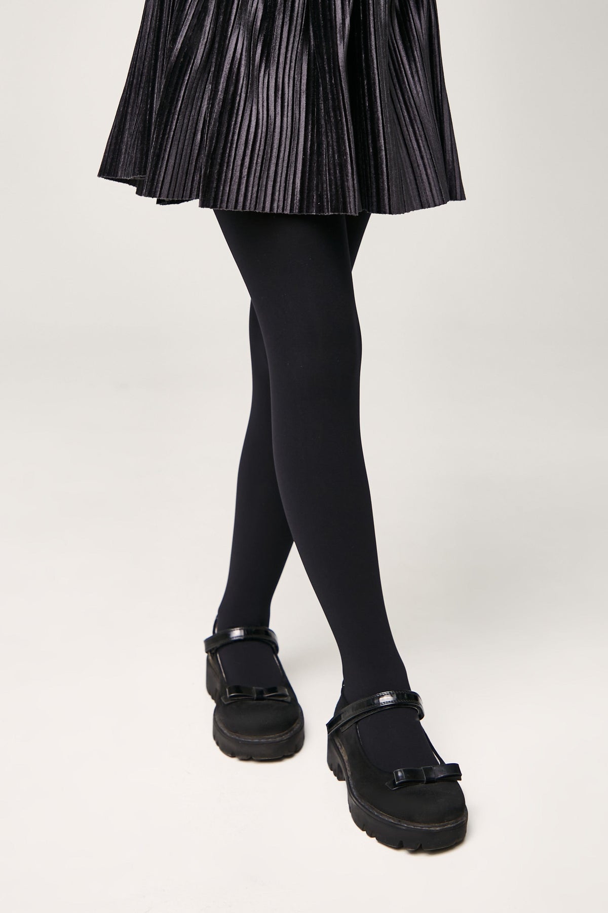 A person wears a pleated black skirt, opaque black tights, and black Mary Jane shoes with bow accents and chunky lug soles.