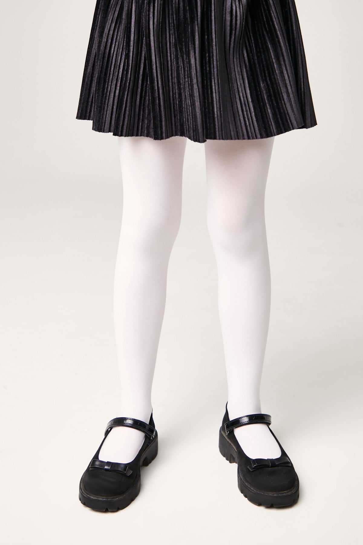 A person wears a pleated black skirt, opaque white tights, and black Mary Jane shoes with buckled straps and small bow accents.