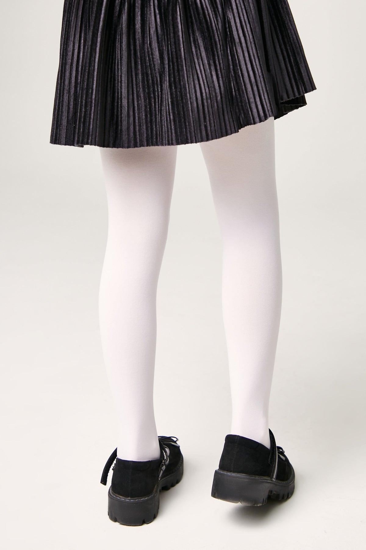 A person wears a pleated black skirt, opaque white tights, and black shoes with side straps.