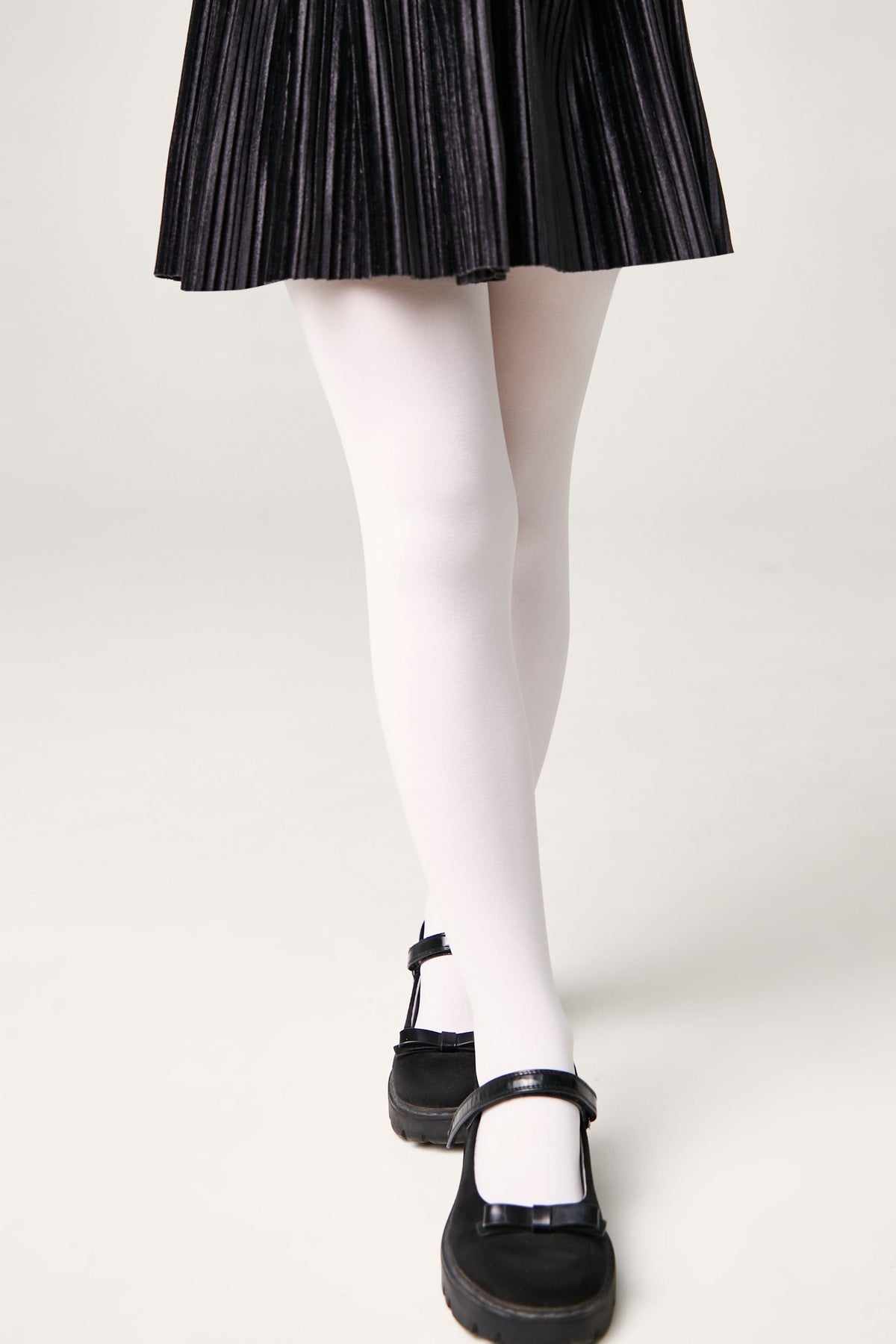 A person wears white opaque tights with a black pleated skirt and black Mary Jane shoes against a light gray background.