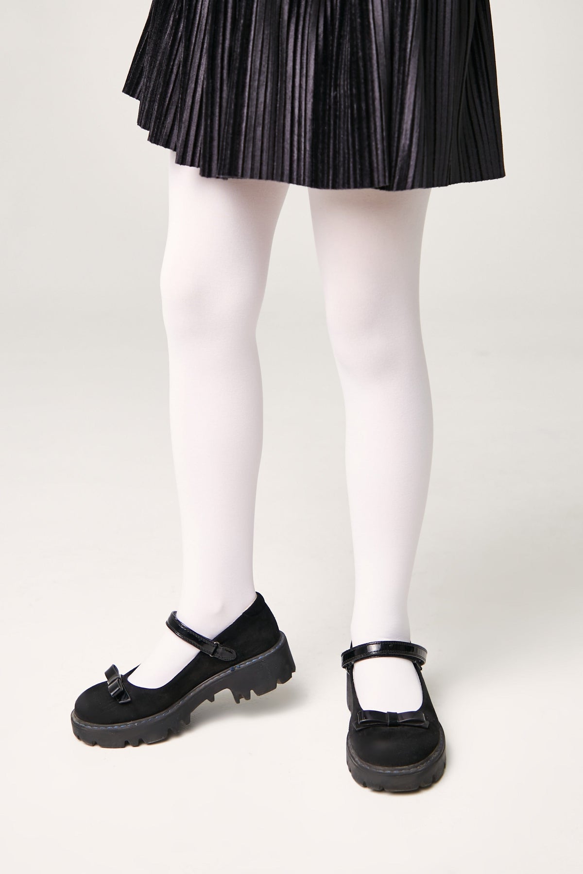 Black mary jane shoes with a chunky lug sole and a bow accent on the strap, worn with white opaque tights and a pleated black skirt.