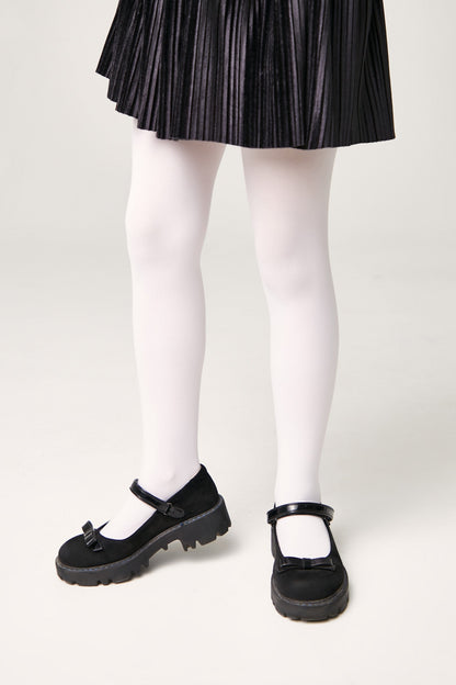 Black mary jane shoes with a chunky lug sole and a bow accent on the strap, worn with white opaque tights and a pleated black skirt.
