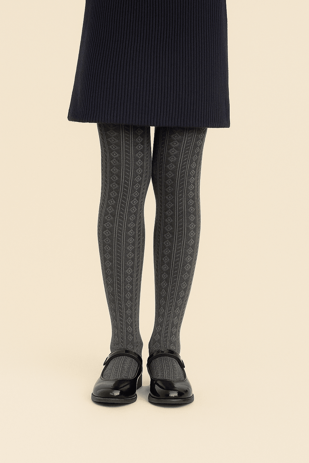 A pair of grey patterned tights featuring vertical lines and diamond shapes worn with a dark navy ribbed skirt and black patent leather Mary Jane shoes against a light beige background.