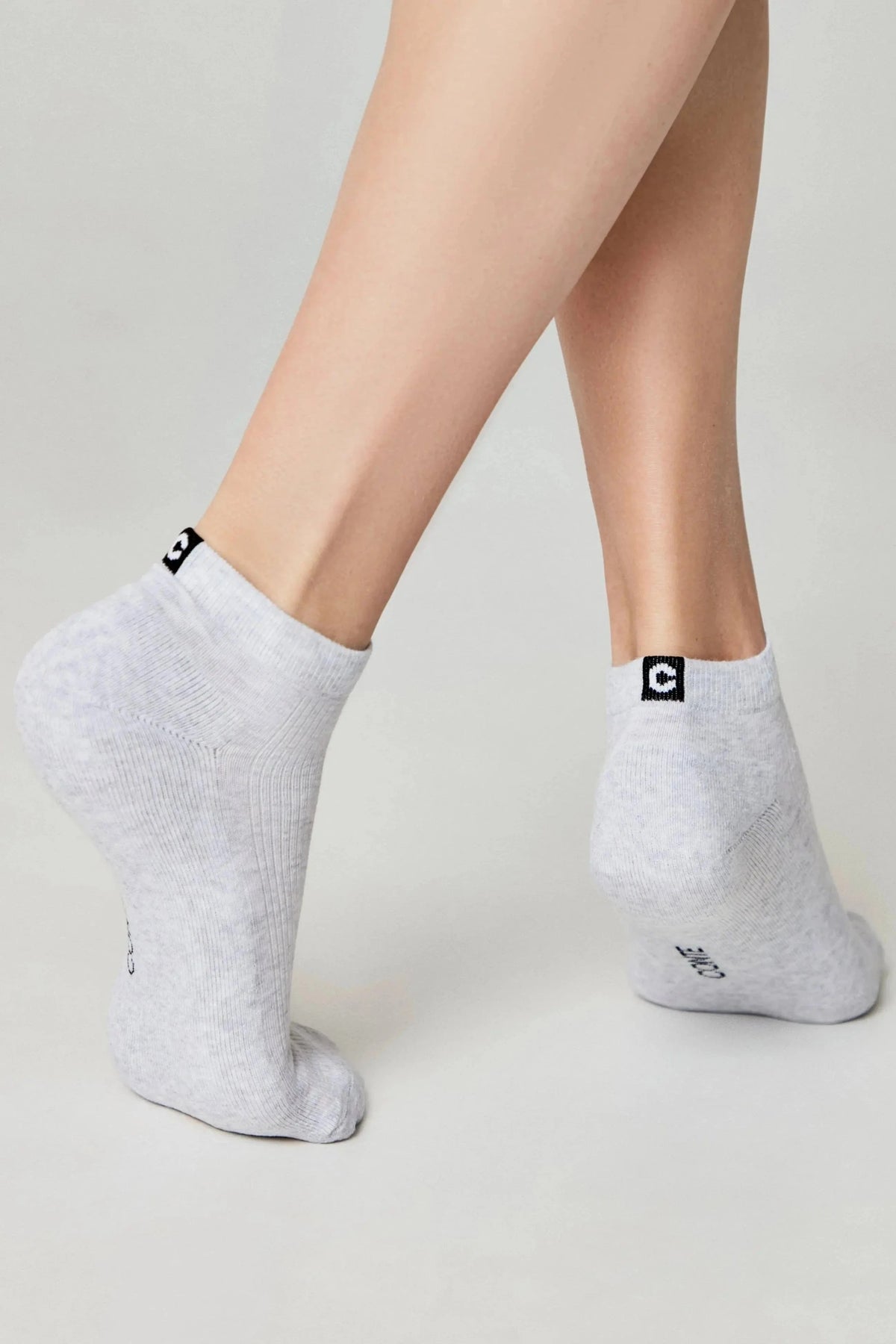 A pair of light gray ankle socks with a black logo patch at the back heel, worn on the feet of a model against a plain light background.