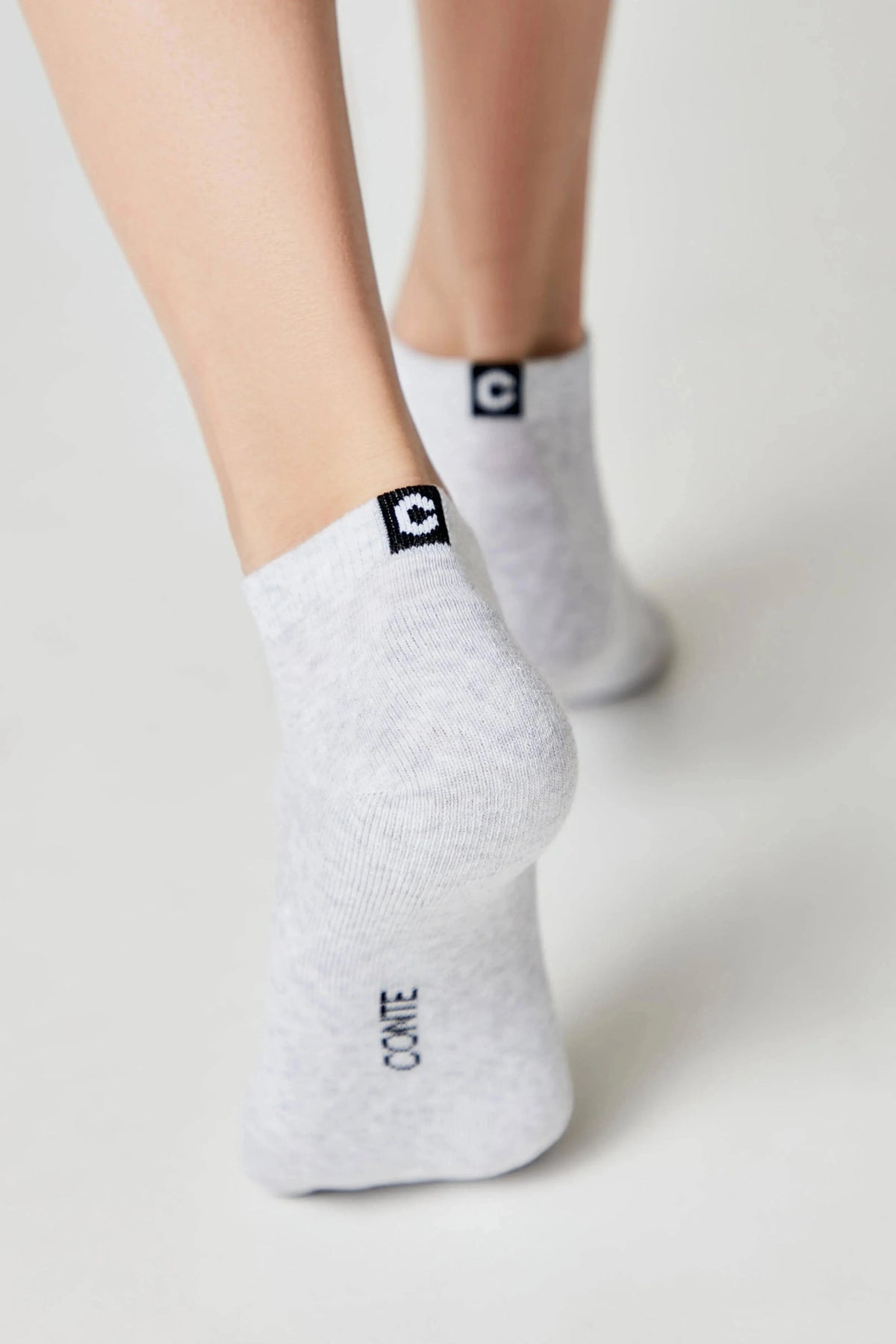 Light grey ankle socks with a black logo patch on the heel and the word CONTE printed on the lower foot, worn on a person's feet against a plain white background.