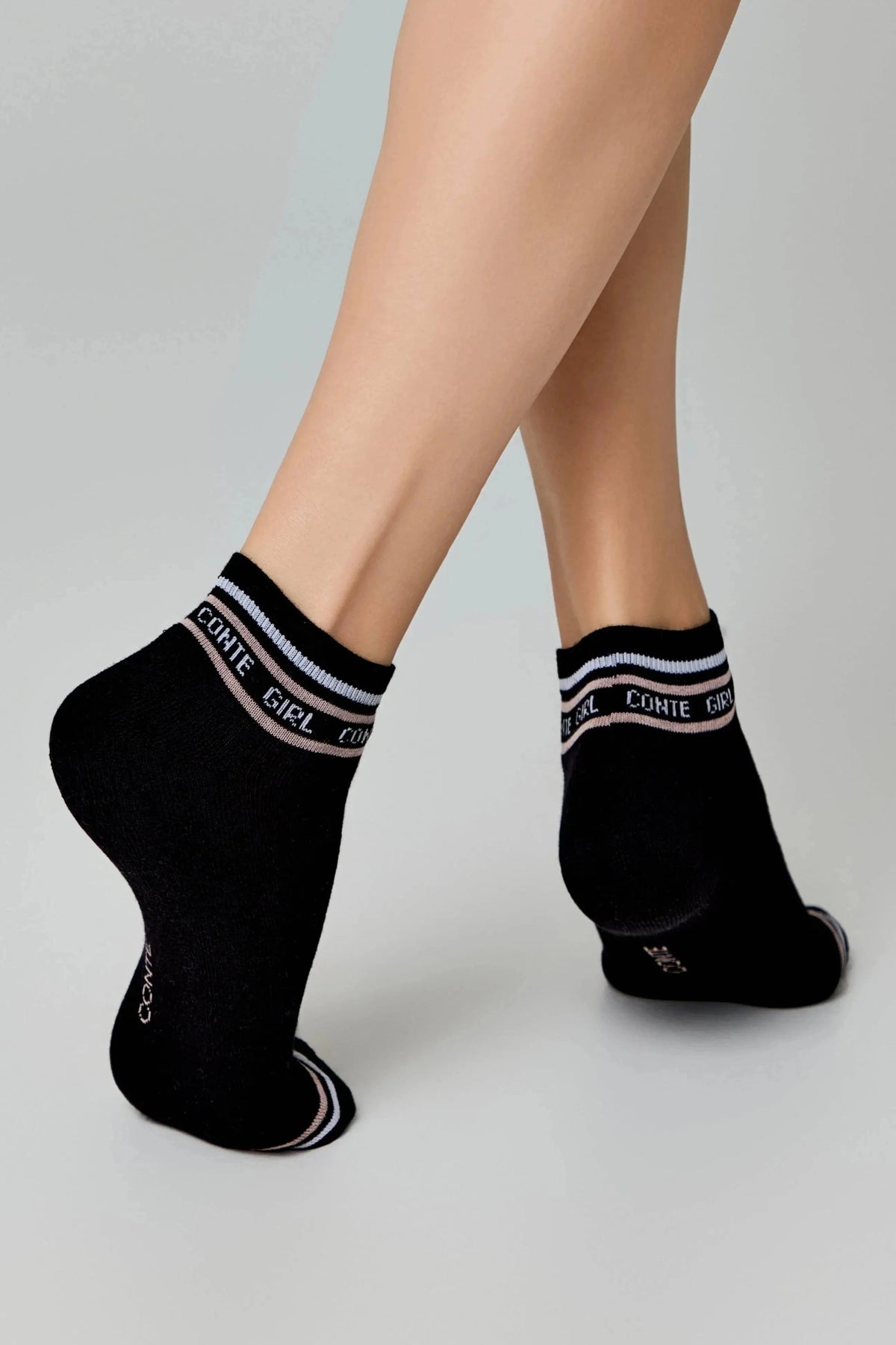 A pair of black ankle socks with white and pink striped trim and the words CONTE GIRL written in white lettering on the cuff, worn on a model's feet against a plain grey background.