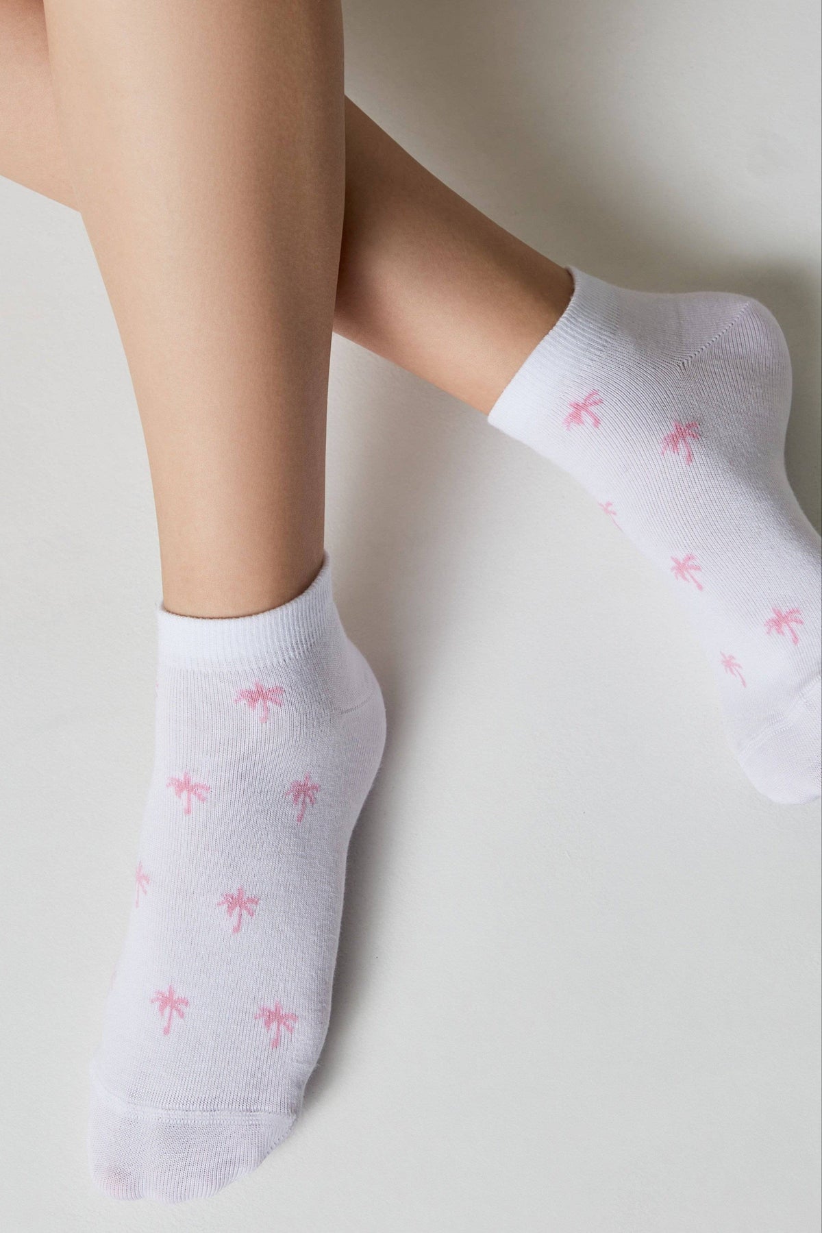 White ankle socks featuring a repeated pattern of small pink palm tree icons worn on a person's feet.