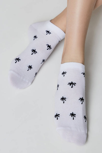 A pair of white ankle socks patterned with small black palm trees worn on a person's feet.