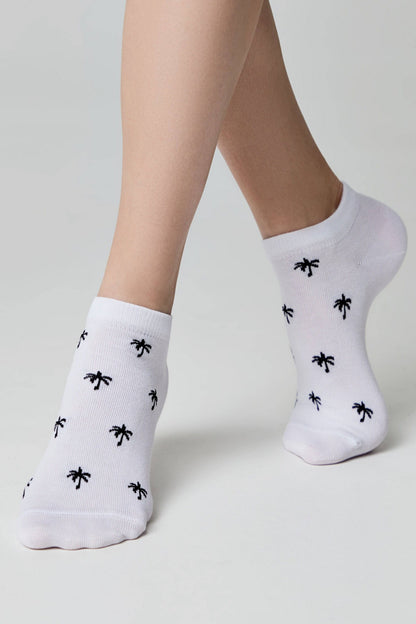 White ankle socks with a repeating small black palm tree pattern worn on feet against a light grey background.