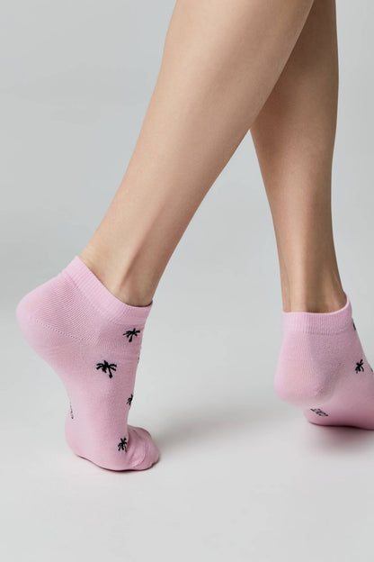A pair of light pink ankle socks featuring a repeated black palm tree pattern, worn on a person's feet against a light grey background.