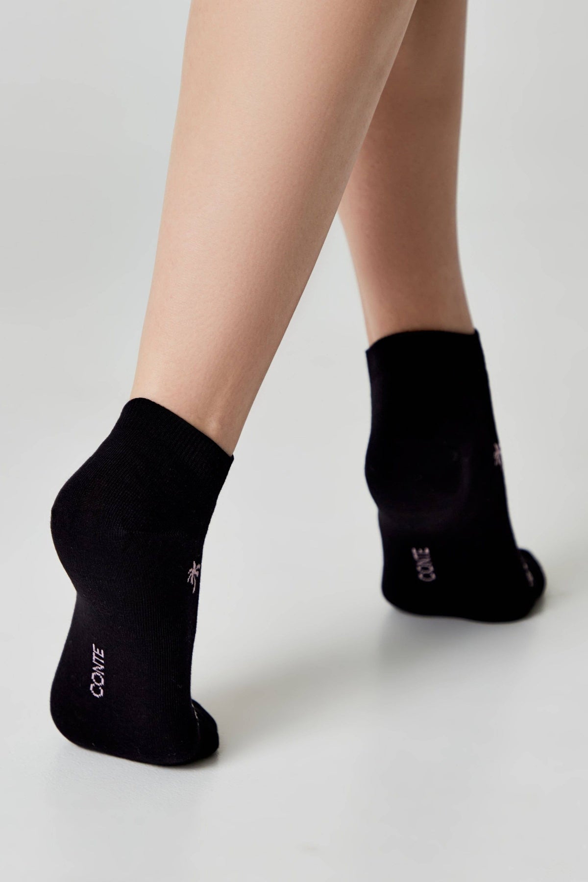 A pair of black ankle socks worn on the feet, featuring the word CONTE and a small palm tree icon embroidered in white on the heel.