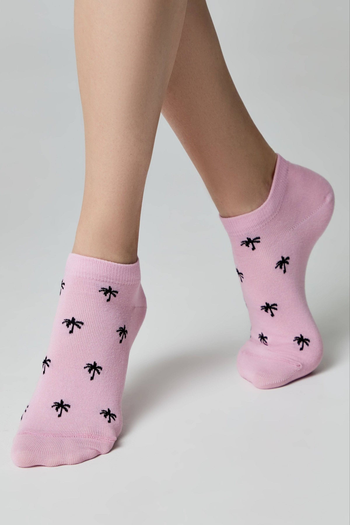 A pair of ankle socks in light pink featuring a repeating pattern of small black palm trees on the wearer's feet.