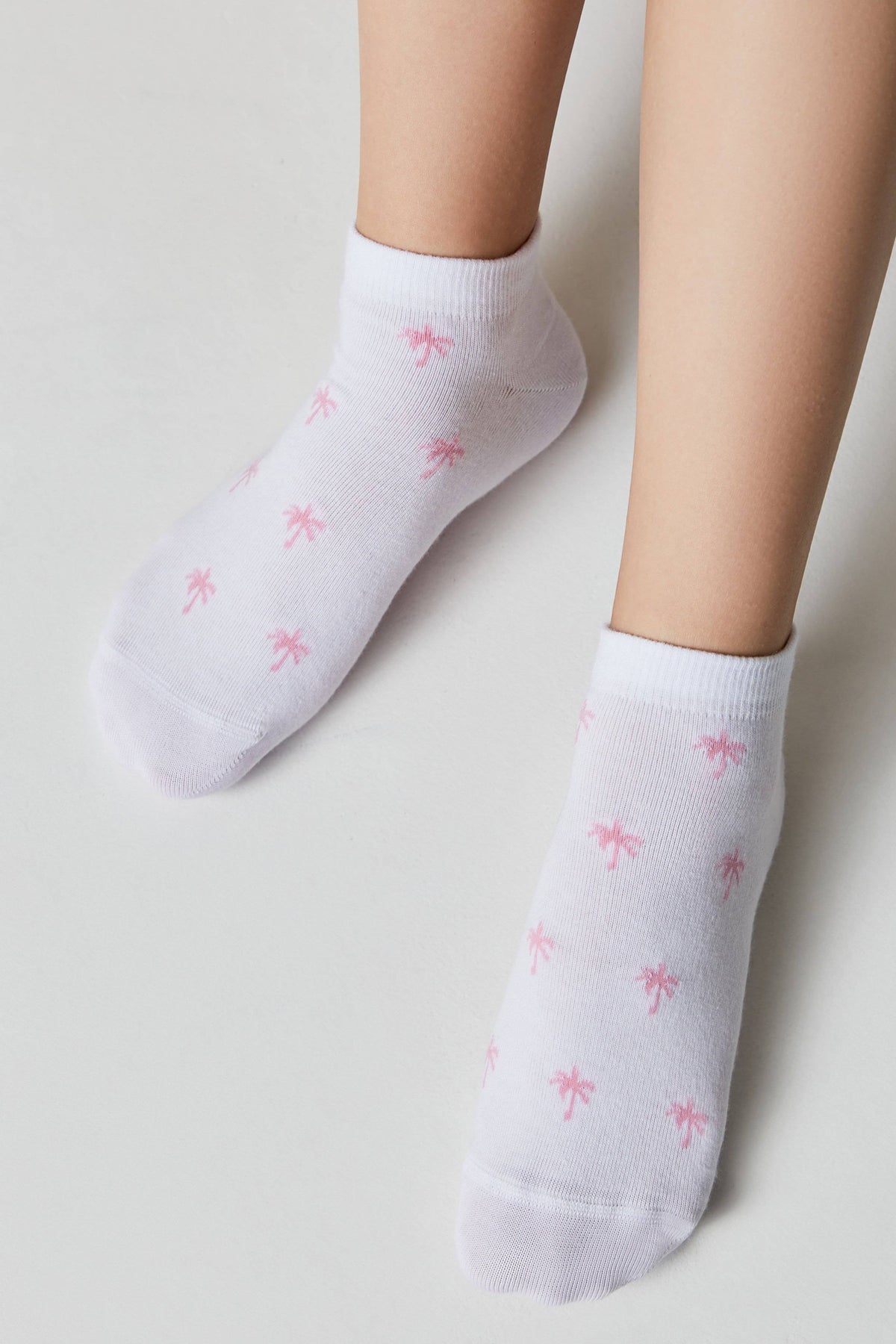 Ankle socks in white with a repeating small pink palm tree print worn on a pair of legs against a plain background.