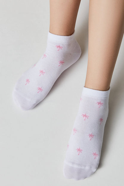 Ankle socks in white with a repeating small pink palm tree print worn on a pair of legs against a plain background.