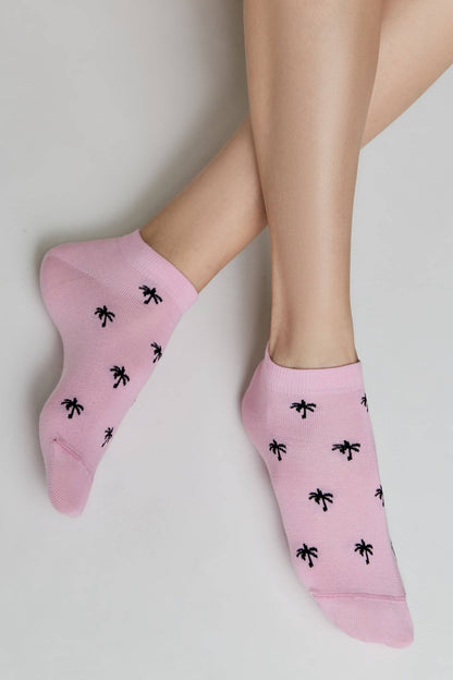 A pair of pink ankle socks with a repeating black palm tree pattern worn on feet against a plain light gray background.