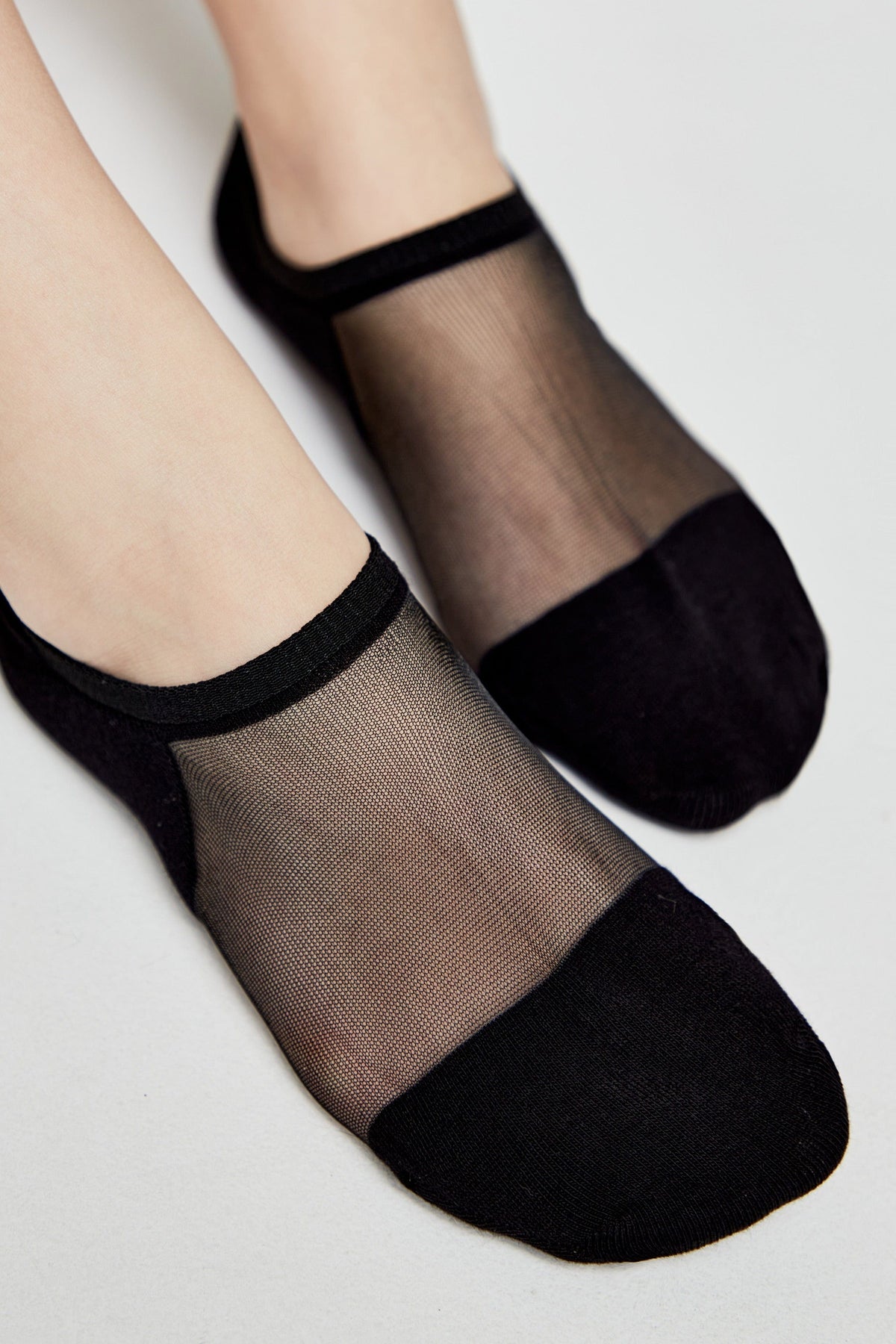 A pair of black no-show socks featuring a sheer mesh top panel and solid black fabric at the toe and heel, shown on a pair of feet against a light grey background.