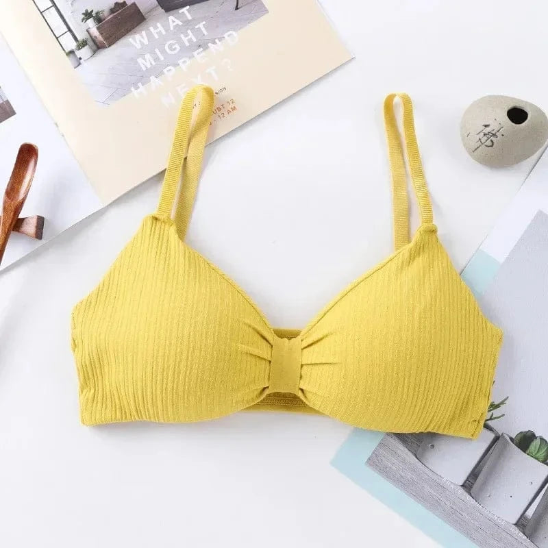 A yellow ribbed bralette with thin shoulder straps and a gathered center detail lays flat on a white surface next to a decorative stone and a printed card.