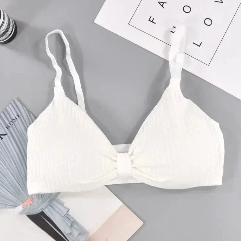 A white bralette with a textured fabric finish and adjustable spaghetti straps, laid flat on a grey surface next to paper stationery.