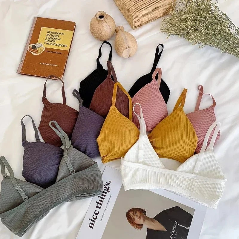 A collection of ribbed, triangular bralettes in various solid colors including white, yellow, gray, purple, brown, pink, and black, displayed on a white surface next to a book and a small vase.