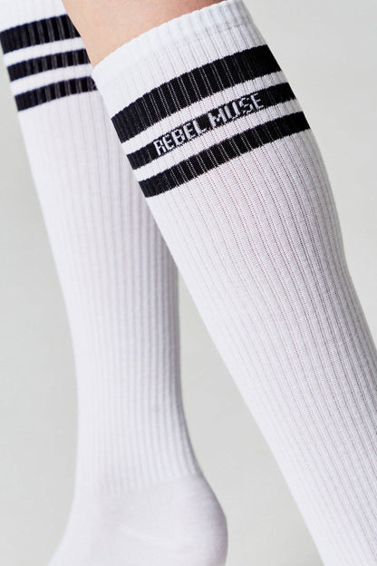 A pair of white knee-high socks featuring two black horizontal stripes and the words REBEL MUSE embroidered in black text below the stripes.