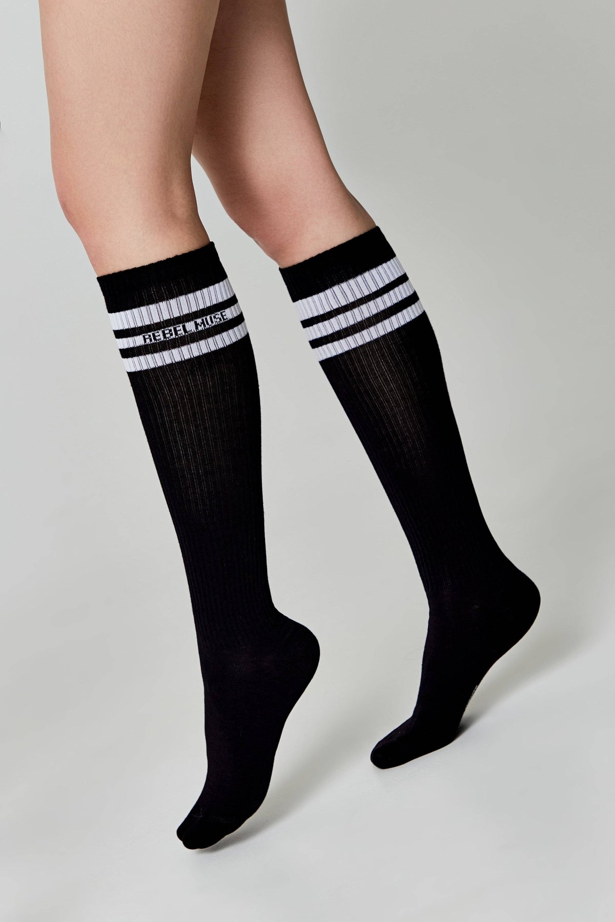 Black knee-high socks feature three white horizontal stripes and white text on the ribbed cuffs, shown worn on a person's legs against a plain light gray background.