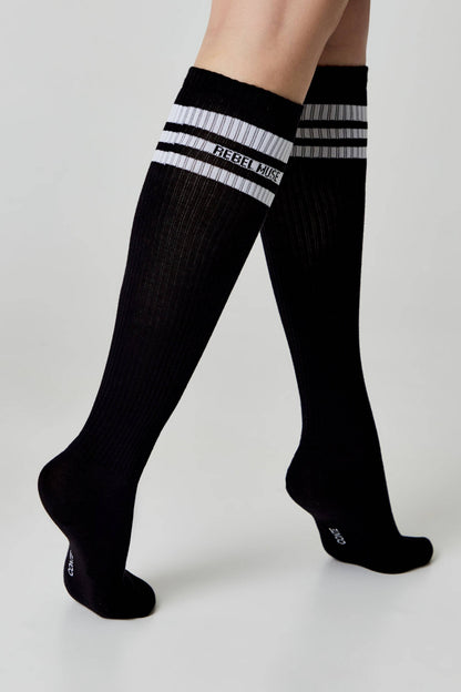 Black knee-high socks feature three white horizontal stripes and the text Rebel Muse printed in white on the ribbed upper cuffs.