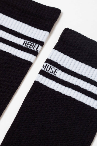 Two black crew socks are laid side-by-side, each featuring two white stripes around the cuff with the word REBEL printed on the left sock and MUSE printed on the right sock.