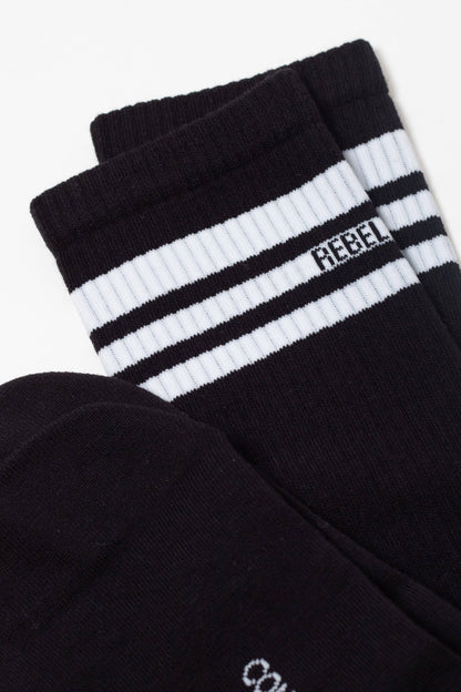 A pair of black crew socks featuring three horizontal white stripes at the top cuff and the word REBEL printed in white lettering.