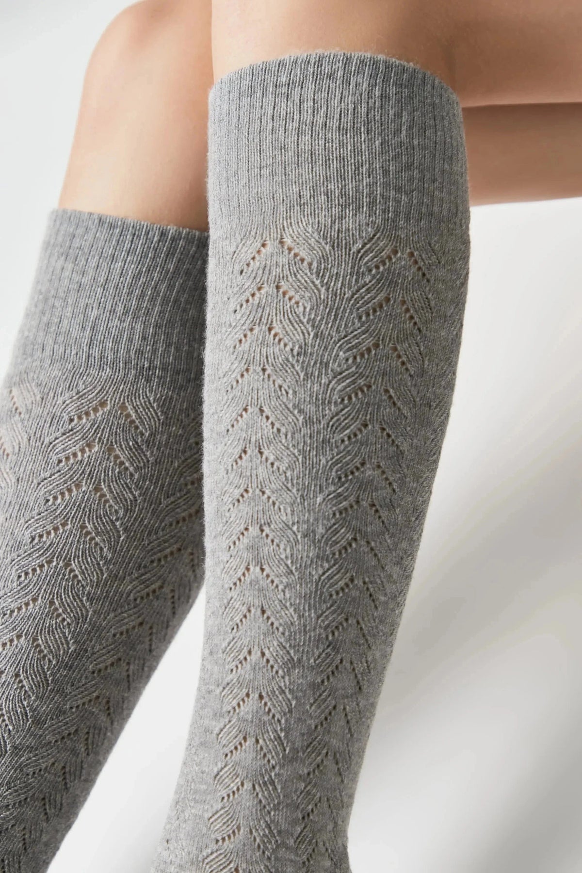 A pair of grey, knee-high socks feature an intricate, vertical openwork leaf pattern against a plain white background.
