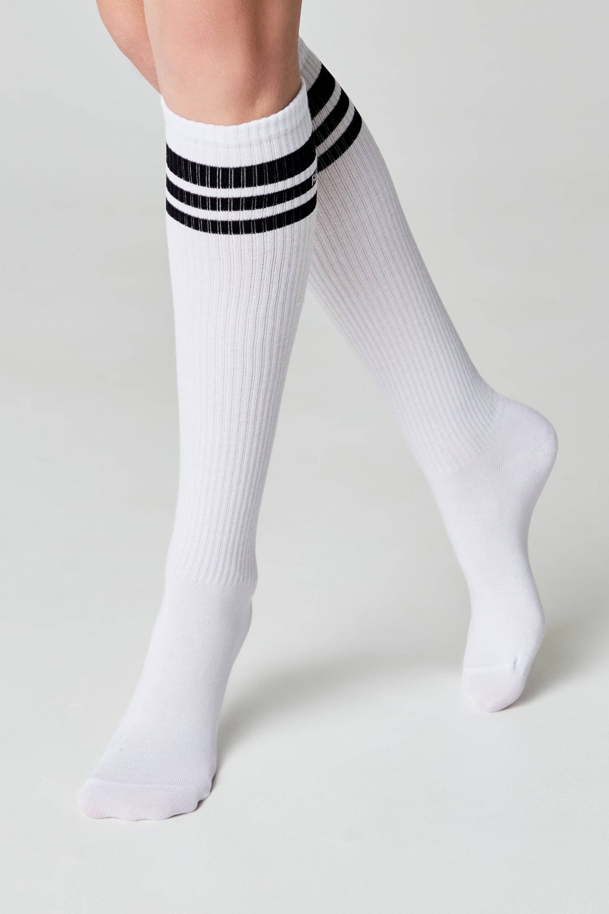 A pair of white knee-high socks featuring three horizontal black stripes at the top, worn on a person's lower legs against a plain light gray background.