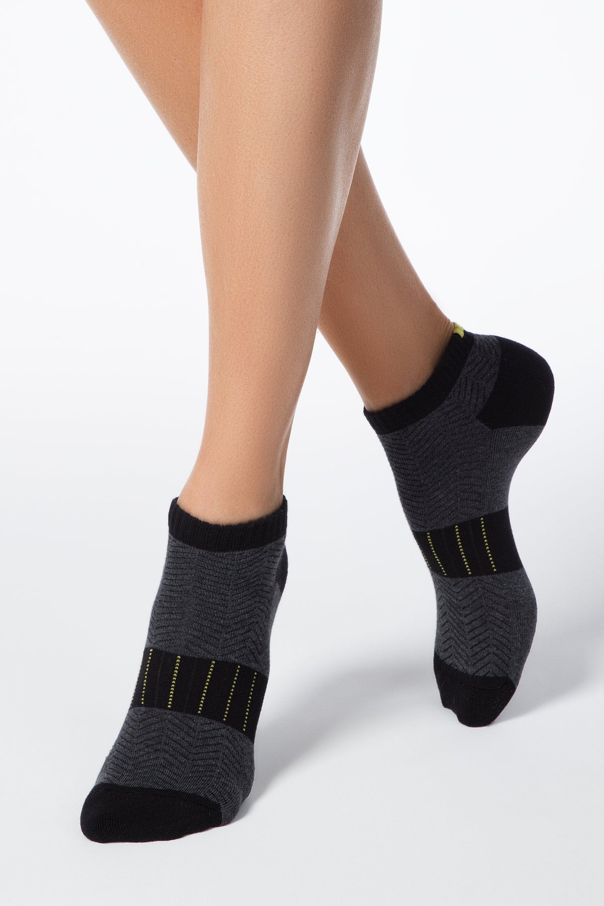 A pair of charcoal gray ankle socks with a black herringbone pattern, black toe and heel sections, and a horizontal band featuring yellow dashed lines across the instep, modeled on crossed legs against a plain white background.